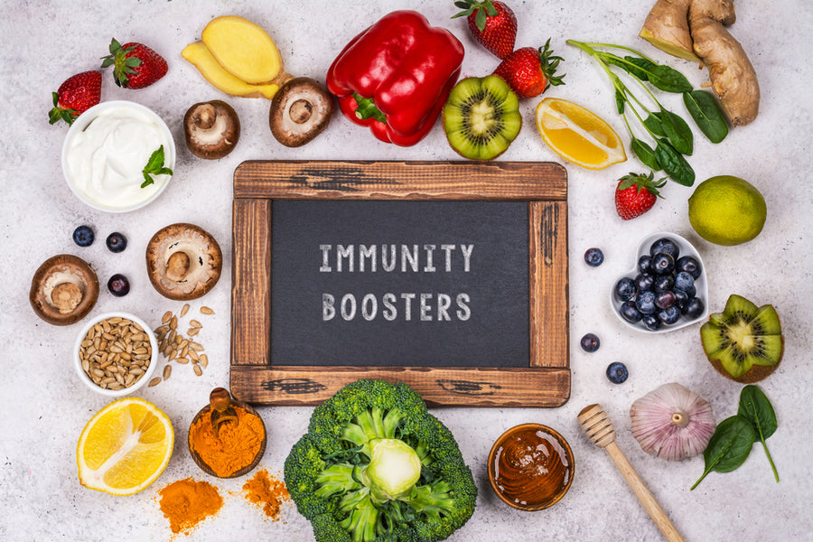 A variety of fresh fruits, vegetables, and spices surround a chalkboard reading "IMMUNITY BOOSTERS," highlighting foods that support immune health.