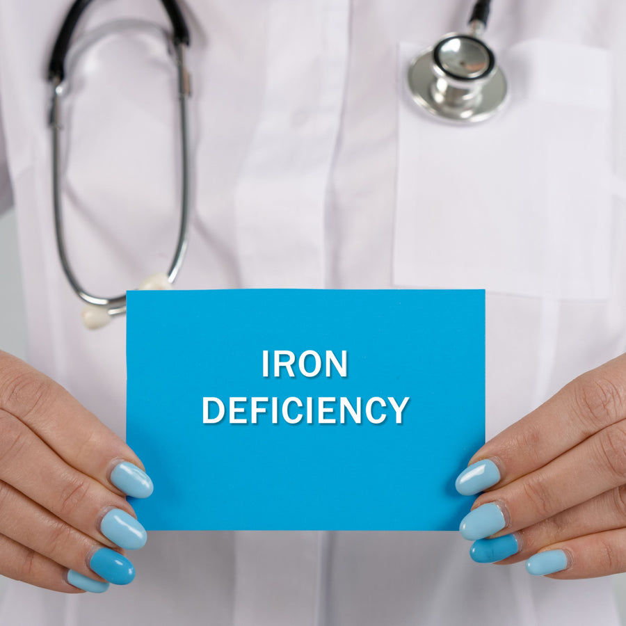 A healthcare professional in a white coat holds a blue card that reads "IRON DEFICIENCY," with a stethoscope visible around their neck, emphasizing the topic of iron-deficiency anemia.