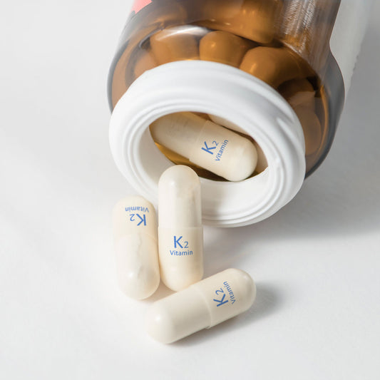 A bottle of vitamin K2 capsules is tipped over, with several white capsules displaying "K2 Vitamin" labels scattered on a white surface.