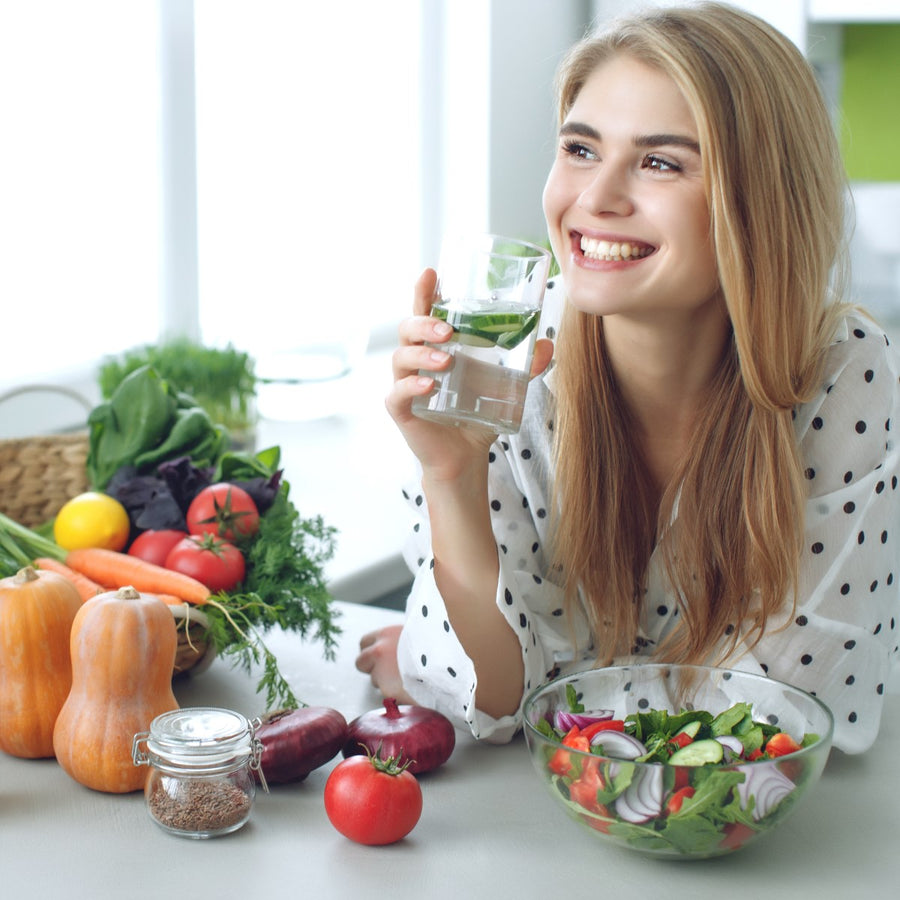 A smiling young woman in a polka dot shirt holds a glass of water with mint, sitting at a table surrounded by fresh vegetables and a bowl of salad in a bright kitchen.