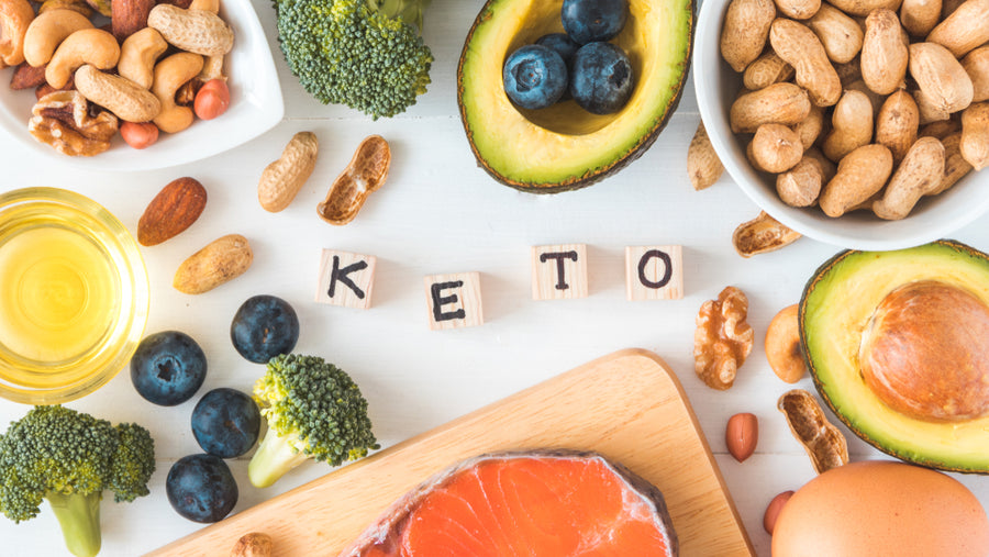 A variety of keto-friendly foods, including broccoli, avocado, nuts, blueberries, and salmon, are arranged on a light surface with wooden blocks spelling "KETO" nearby.