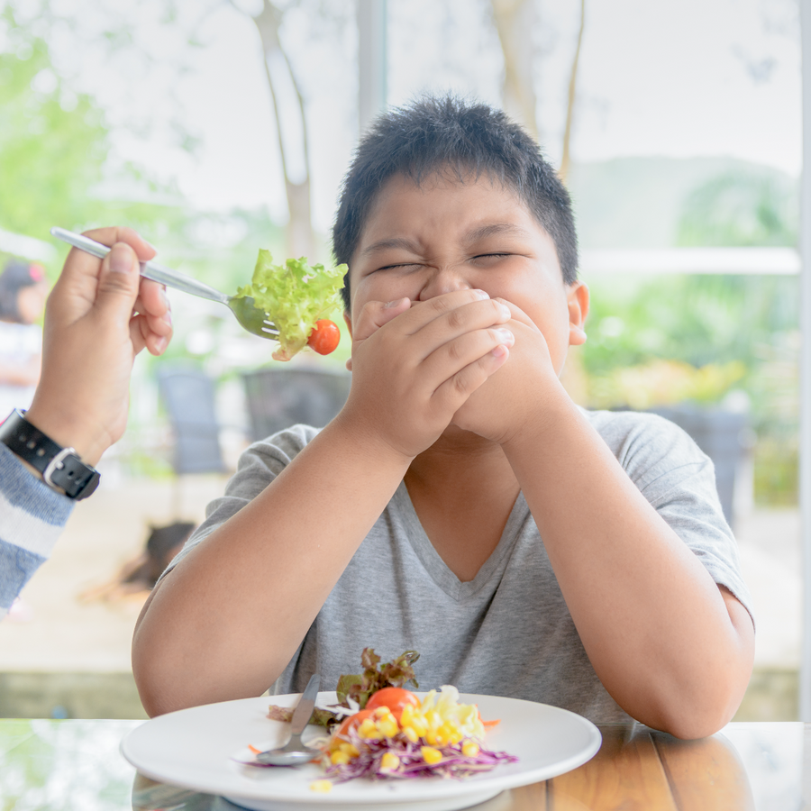 A boy grimaces and covers his mouth while sitting at a table, as someone offers him a forkful of salad in a bright, outdoor dining setting.