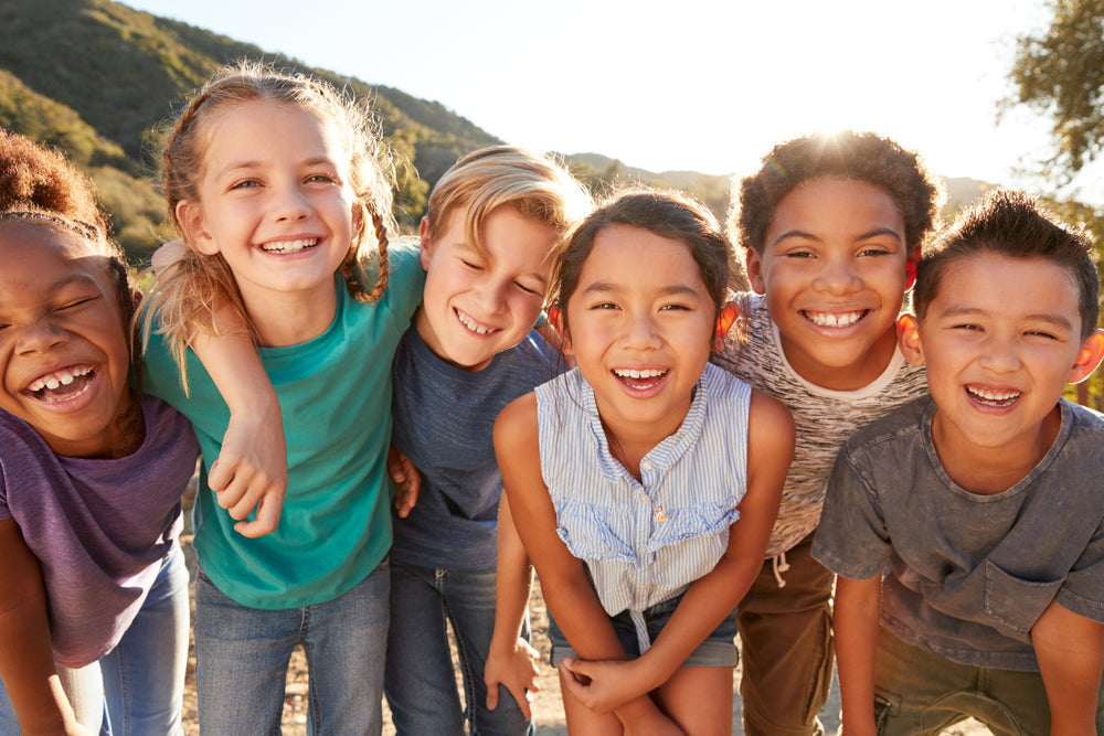 Six smiling children of diverse backgrounds stand closely together outdoors, with a sunny backdrop of hills, expressing joy and friendship while promoting a healthy lifestyle.
