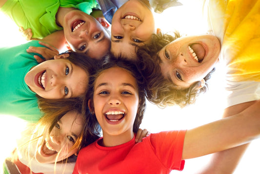 Six smiling children with diverse hairstyles and clothing colors are gathered in a circle, looking down at the camera, radiating joy and friendship in a bright outdoor setting.