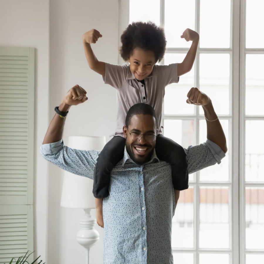 A smiling man lifts a child on his shoulders, both flexing their arms in a playful display of strength, set in a bright room with large windows.