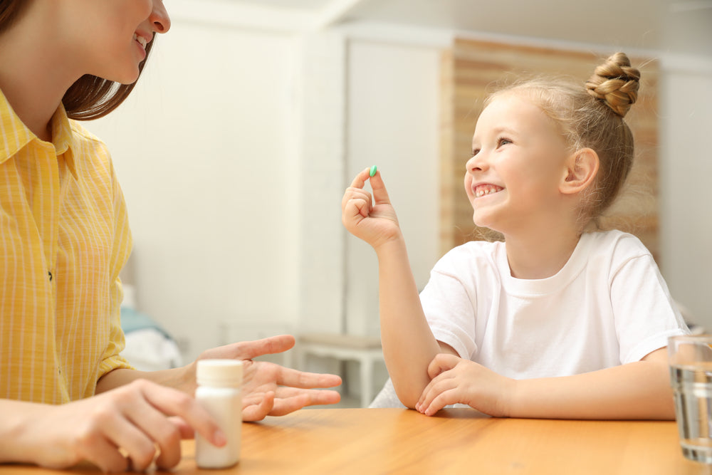 A smiling girl in a white t-shirt holds a green vitamin while sitting at a table, engaging happily with a woman in a yellow shirt, suggesting a positive moment around taking supplements.