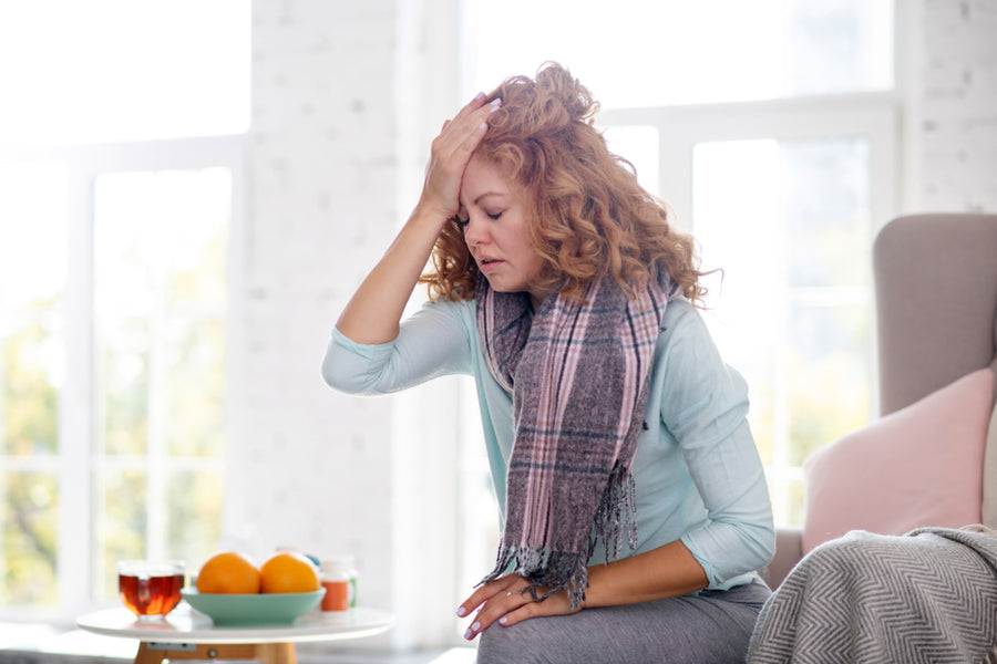A woman with curly red hair sits on a couch, wearing a scarf, looking distressed while holding her forehead, with a table of oranges and tea in a bright, cozy room.