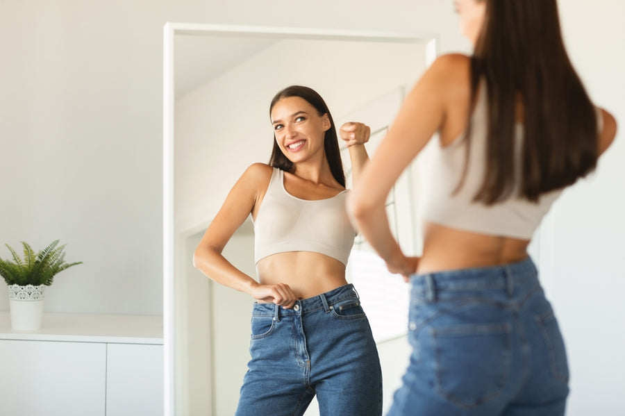 A smiling woman in a fitted tank top and high-waisted jeans admires her reflection in a mirror, showcasing her confidence and satisfaction with her weight loss journey in a bright, modern room.