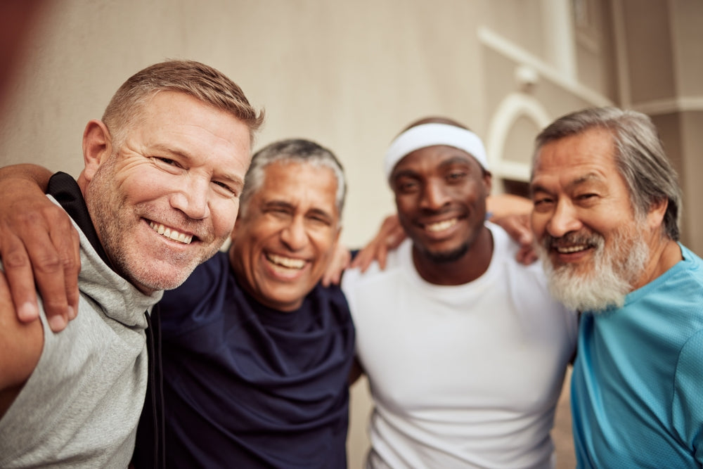Four men of diverse ages and backgrounds smile and pose together with arms around each other in a casual outdoor setting, promoting camaraderie and healthy living.