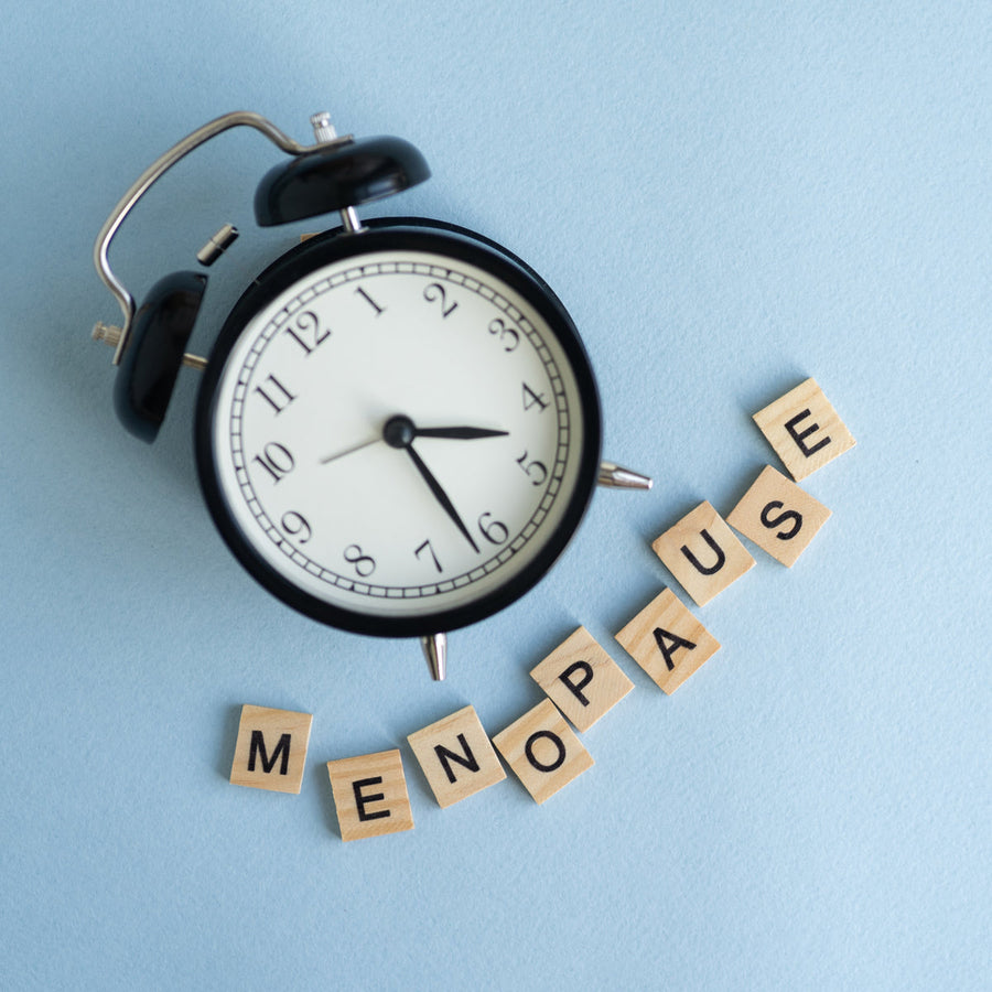 A black alarm clock sits on a light blue background next to wooden letter tiles that spell out "MENOPAUSE," highlighting the theme of time and menopause.