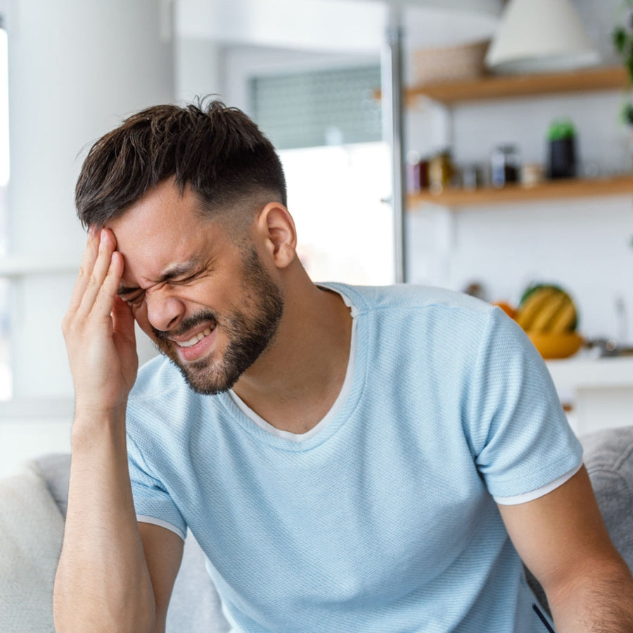 A man with short hair and a beard sits on a couch, grimacing and holding his forehead in pain, in a bright, modern kitchen setting.