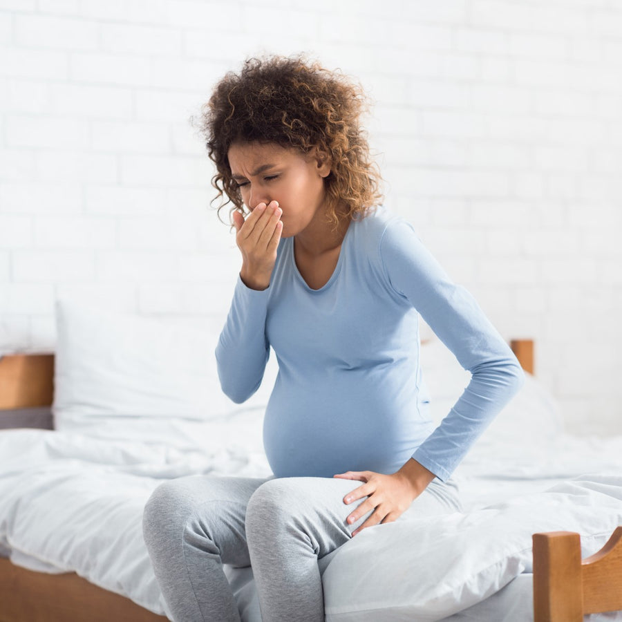 A pregnant woman sits on the edge of a bed, looking distressed and covering her mouth with one hand, in a bright, softly lit room with white walls and bedding.