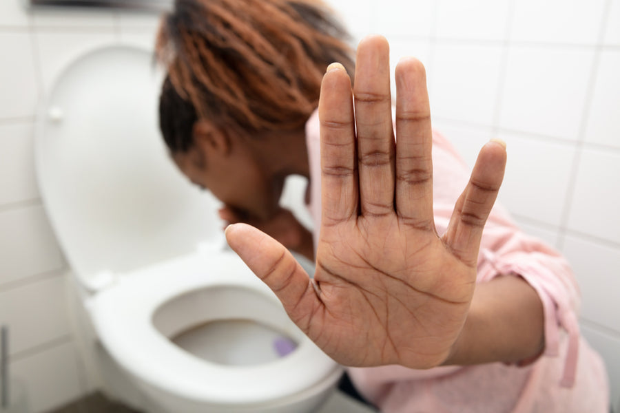 A person with curly hair leans over a toilet, appearing unwell, while holding up a hand to signal for help or to indicate they are not feeling well.