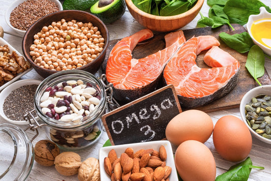 A variety of foods rich in omega-3 fats, including salmon fillets, nuts, seeds, beans, eggs, and leafy greens, are arranged on a wooden surface with a chalkboard labeled "Omega 3."