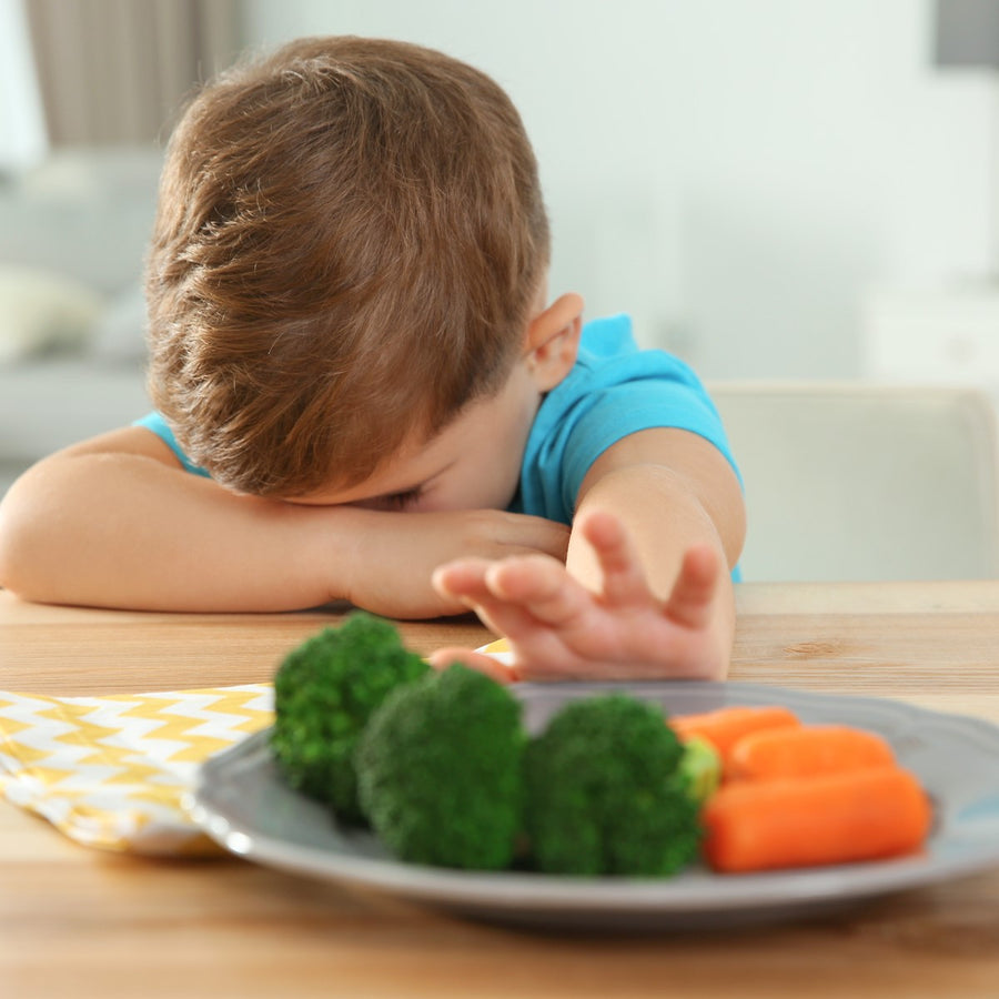 A young boy in a blue shirt rests his head on his arms at a table, looking frustrated as he reaches toward a plate of broccoli and carrots.