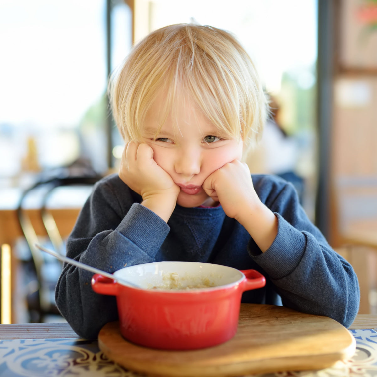 A young child with blonde hair sits at a table, resting their chin on their hands, looking displeased while staring at a small bowl of food in a cozy dining setting.