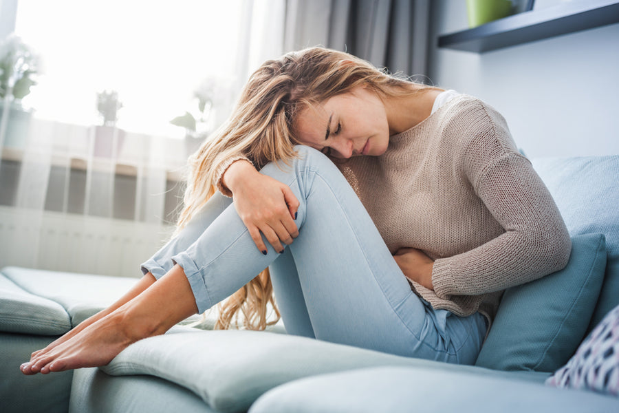 A woman sits on a couch, hugging her knees and resting her forehead on them, looking pained, in a bright room with plants, suggesting discomfort related to PMS symptoms.
