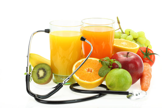 Two glasses of juice sit beside a variety of fresh fruits and vegetables, including bananas, oranges, and grapes, with a stethoscope placed among them, symbolizing the connection between nutrition and health.