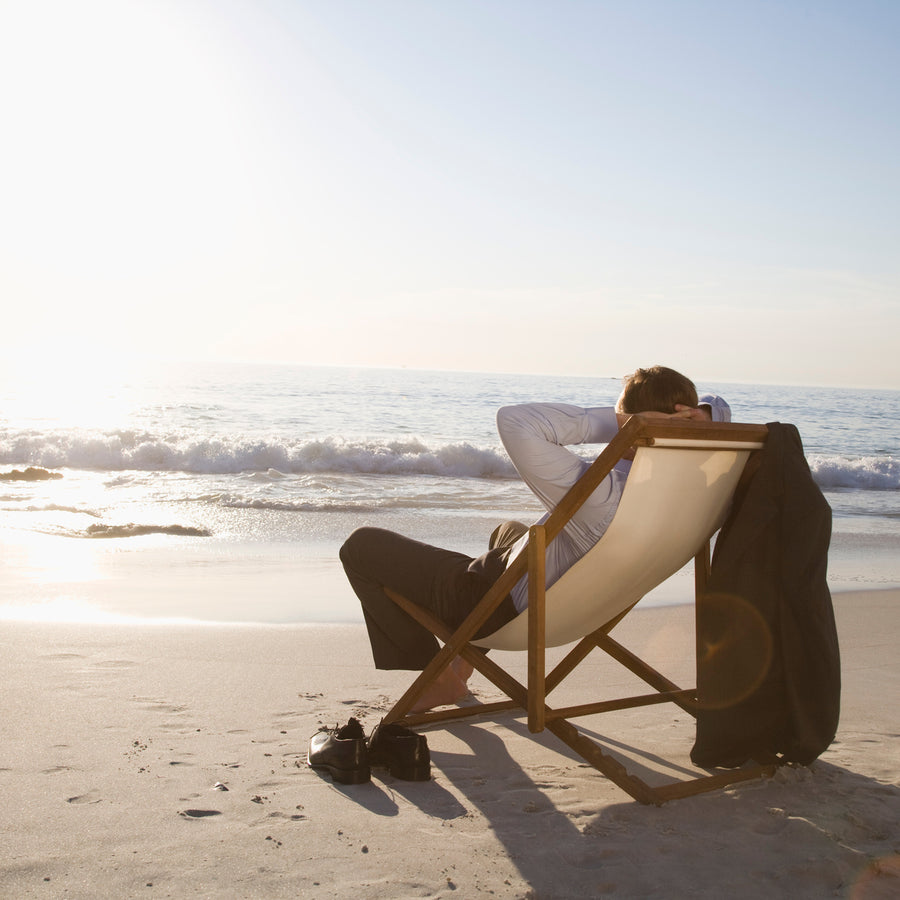 A person in a suit relaxes in a beach chair, leaning back with hands behind their head, as gentle waves lap at the shore under a bright sky.