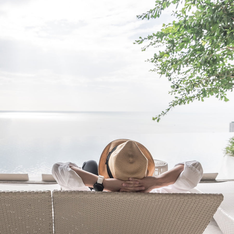 A person wearing a straw hat and white shirt relaxes on a lounge chair with arms behind their head, gazing at a tranquil ocean view under a cloudy sky.