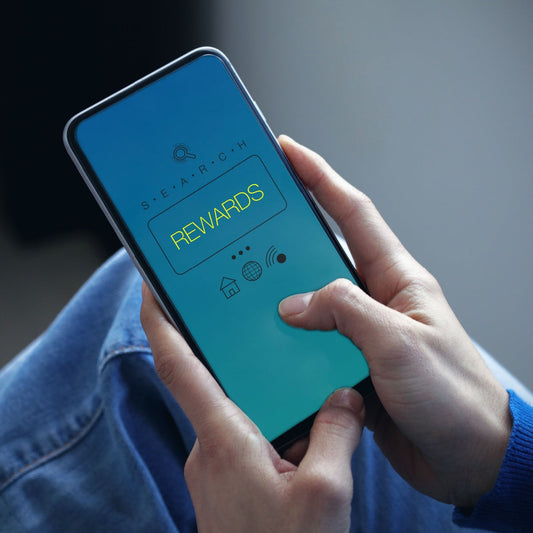 A person holds a smartphone displaying a blue gradient screen with the word "REWARDS" in bright yellow, indicating they are engaged in a search for rewards on the device.