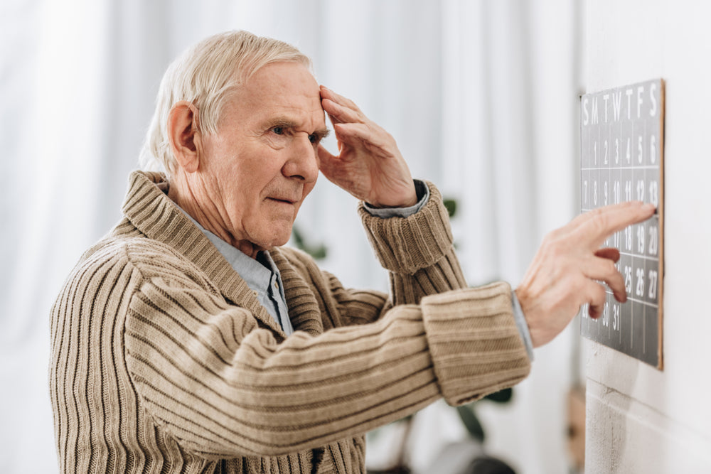 An elderly man in a cozy sweater touches his forehead in concentration while pointing at a calendar on the wall, reflecting a struggle with memory in a well-lit indoor setting.