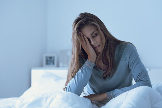 A woman sits on a bed, looking tired and distressed, with her hand on her forehead, in a softly lit bedroom, reflecting the struggles of dealing with vitamin C deficiency symptoms.