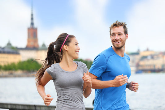 A woman and a man jog together along a waterfront, smiling and enjoying their exercise, with a city skyline and a church tower visible in the background.
