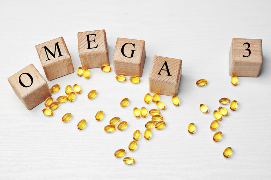 Wooden blocks spelling "OMEGA" and "3" are arranged on a white surface, surrounded by scattered yellow omega-3 gel capsules, highlighting the topic of omega-3 vitamin patches.