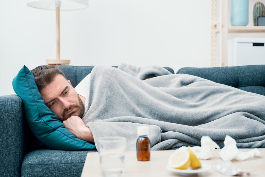 A man lies on a couch wrapped in a blanket, looking tired and contemplative, with a glass of water, a bottle of medicine, tissues, and lemon on a table nearby in a bright, modern living room.