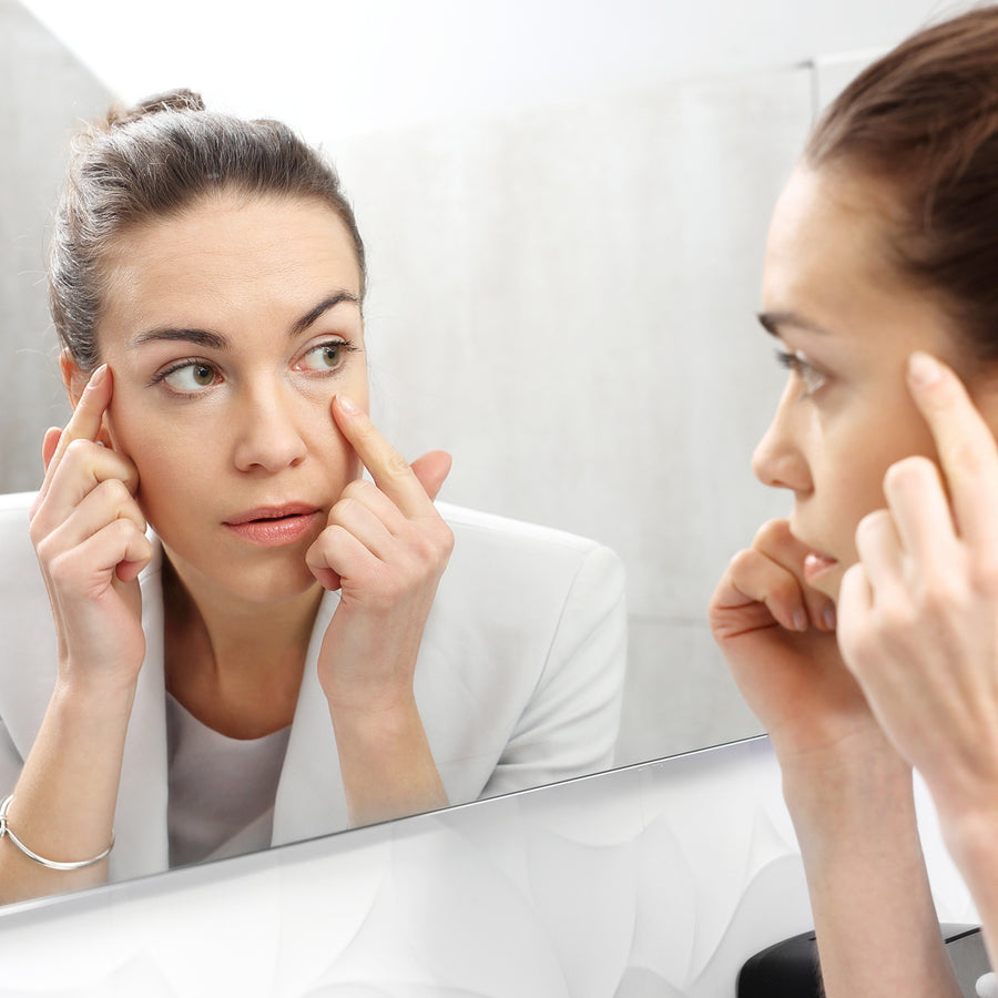 A woman in a white blazer examines her reflection in a mirror, gently pressing her fingers against the skin around her eyes, focusing on reducing signs of aging.