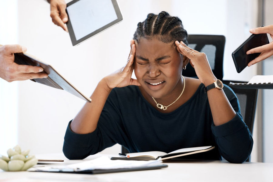A woman with braided hair sits at a table, visibly stressed with her hands on her temples, while two hands offer her devices, highlighting the pressure of multitasking in a modern setting.
