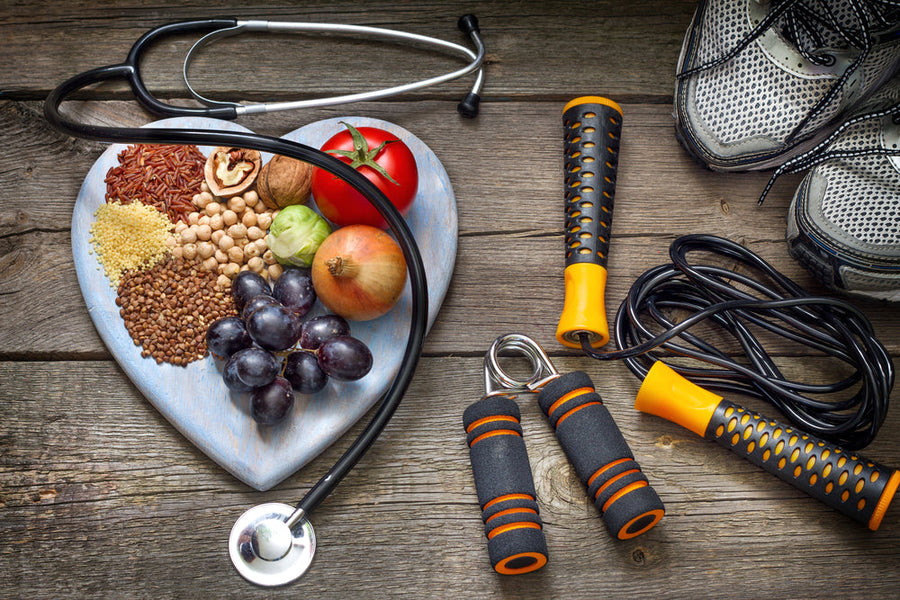 A heart-shaped wooden plate holds various healthy foods like grains, fruits, and vegetables, alongside a stethoscope, workout equipment, and running shoes on a rustic wooden surface.