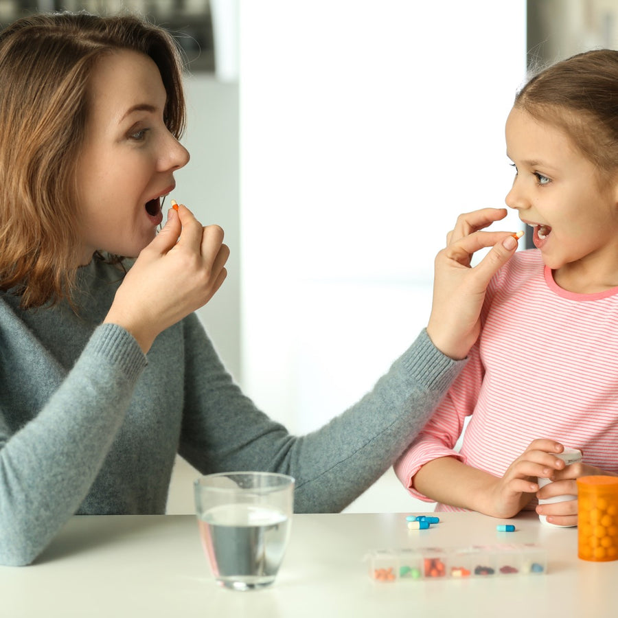 A woman and a girl sit at a table, both smiling as the woman holds a pill while the girl prepares to take one, with a glass of water and a container of pills nearby.