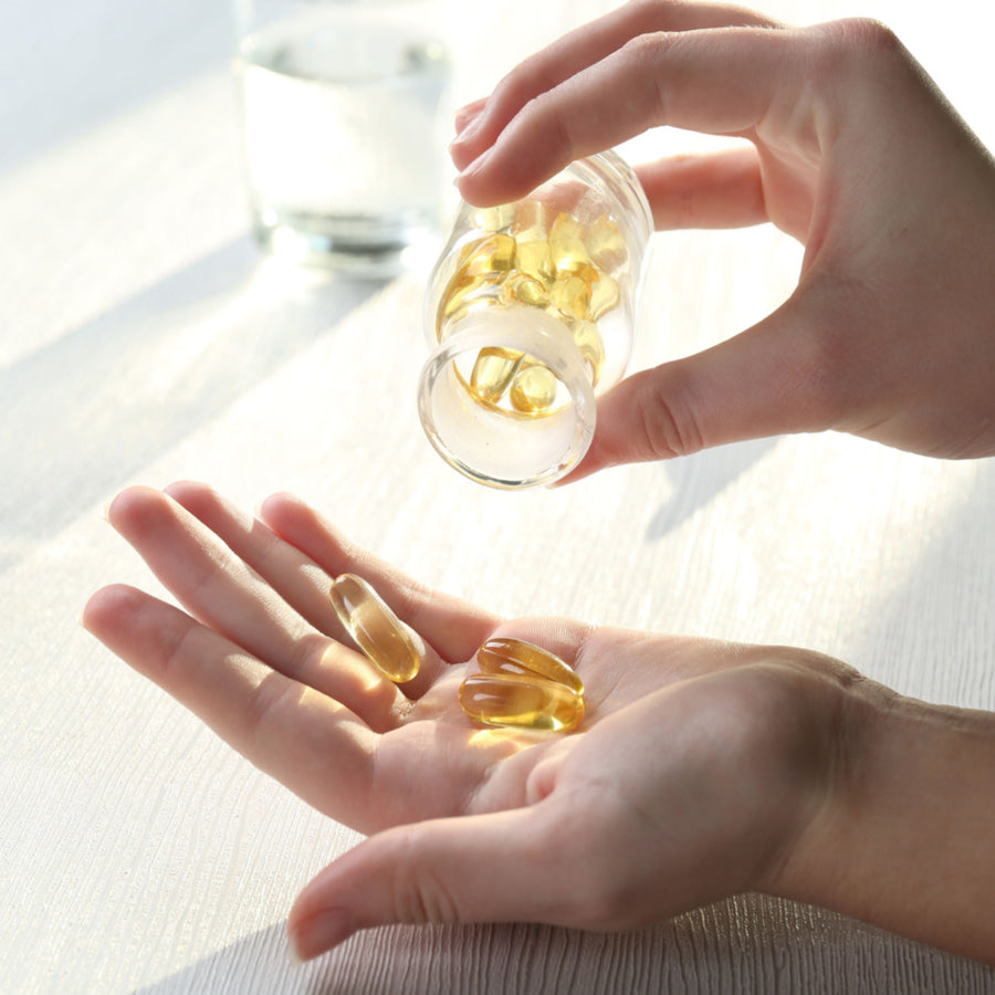 A hand holds a glass jar pouring golden vitamin capsules into an open palm, with a glass of water in the background on a light wooden surface.