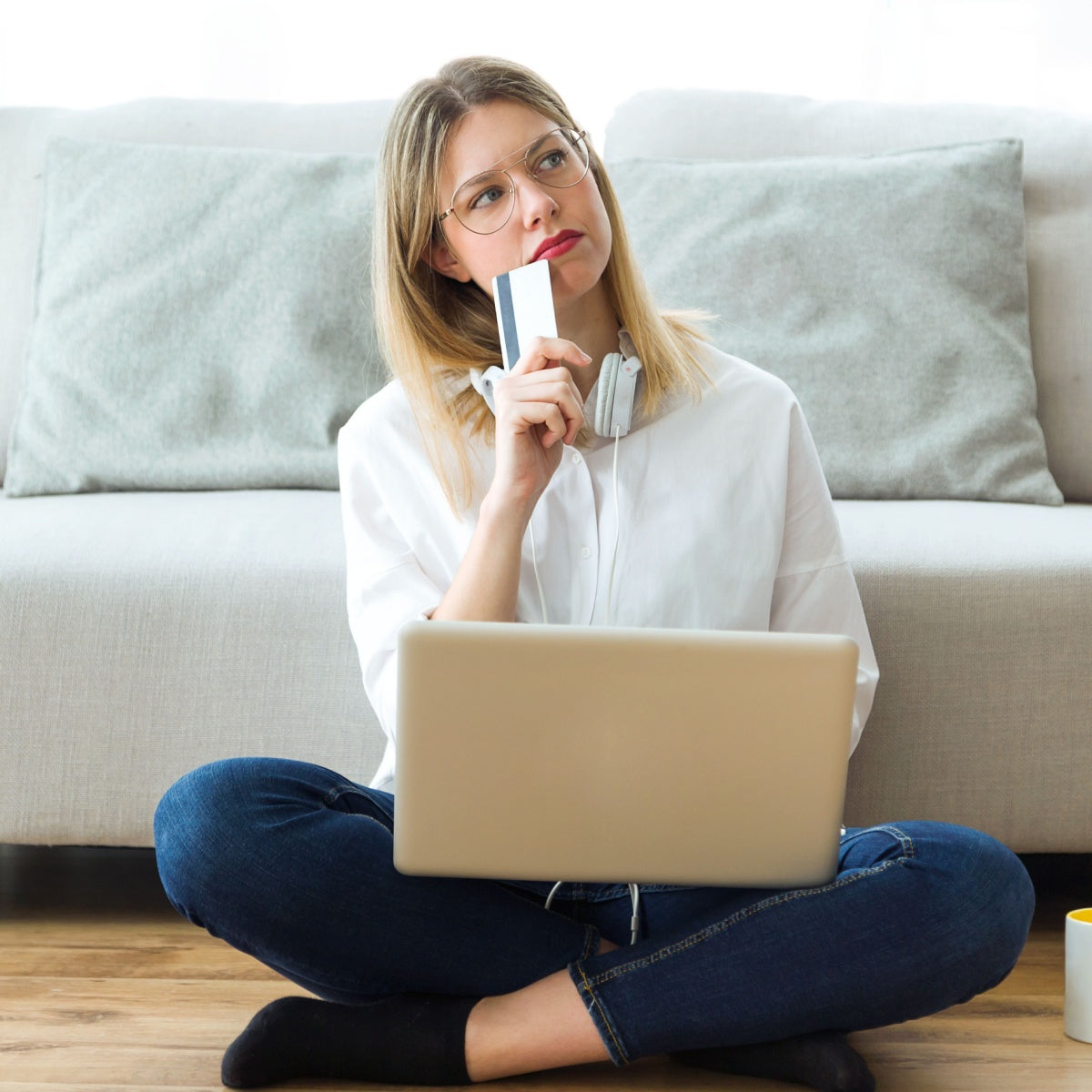 A woman sits cross-legged on the floor with a laptop in front of her, holding a credit card and looking thoughtfully, wearing headphones and a white shirt, with a sofa and a mug nearby.