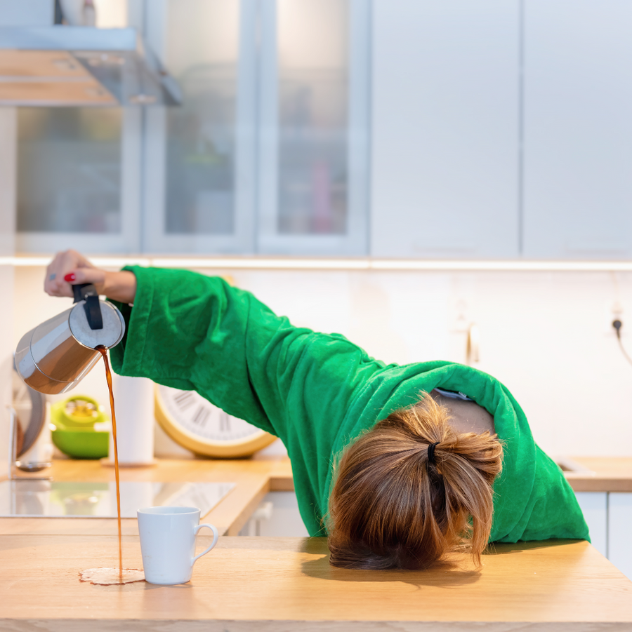 A person in a green robe leans over a kitchen counter, pouring coffee from a French press into a white mug, with spilled coffee pooling on the wooden surface.