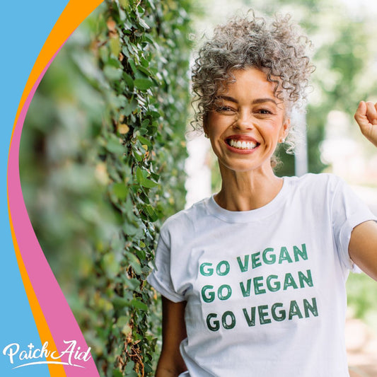 A smiling woman with curly hair raises her fist in a leafy outdoor setting, wearing a white t-shirt that says "GO VEGAN" multiple times, promoting a vegan lifestyle.