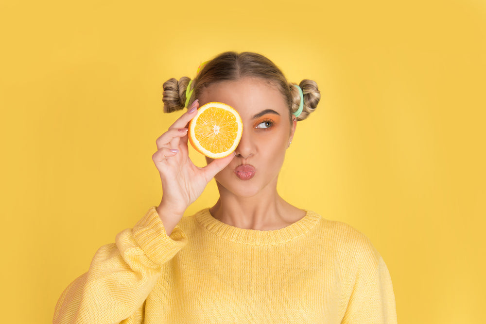 A woman in a yellow sweater playfully holds a halved orange to her face, making a playful expression against a bright yellow background, emphasizing the theme of vitamin C and wellness.