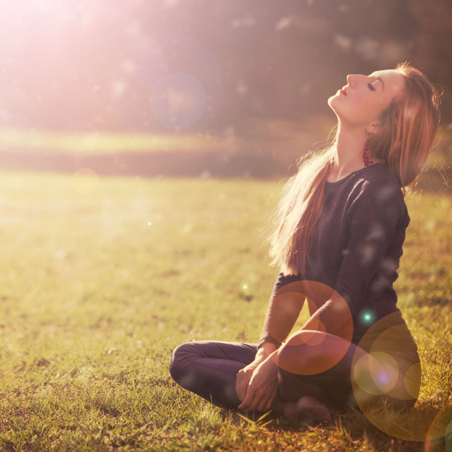 A woman sits cross-legged on green grass, tilting her head back with closed eyes, basking in warm sunlight, conveying a sense of peace and relaxation, highlighting the benefits of Vitamin D.