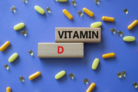 Wooden blocks spelling "VITAMIN" and "D" are centered on a purple background, surrounded by various vitamin capsules in yellow, green, and clear colors, emphasizing the importance of Vitamin D.