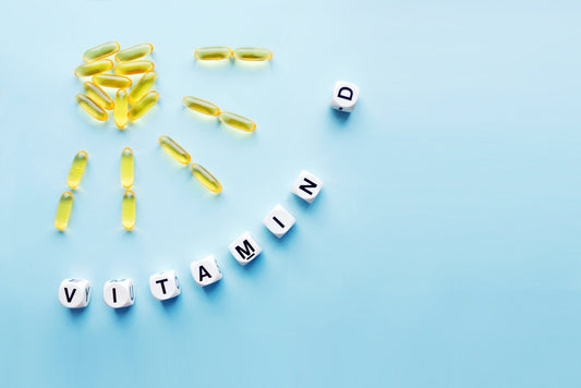 Yellow gelatin capsules are arranged in a sun shape, with white letter blocks spelling "VITAMIN D" on a light blue background, highlighting the importance of vitamin D for health.