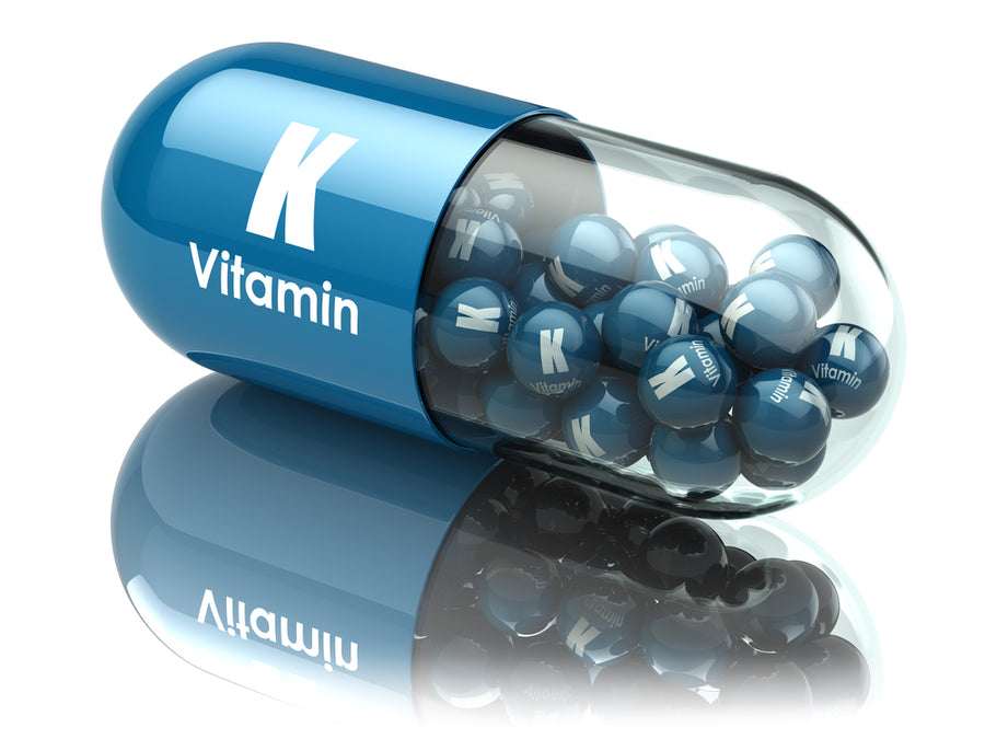 A blue capsule labeled "Vitamin K" contains shiny blue balls with the same label, reflecting light on a smooth surface, highlighting the importance of this vitamin.
