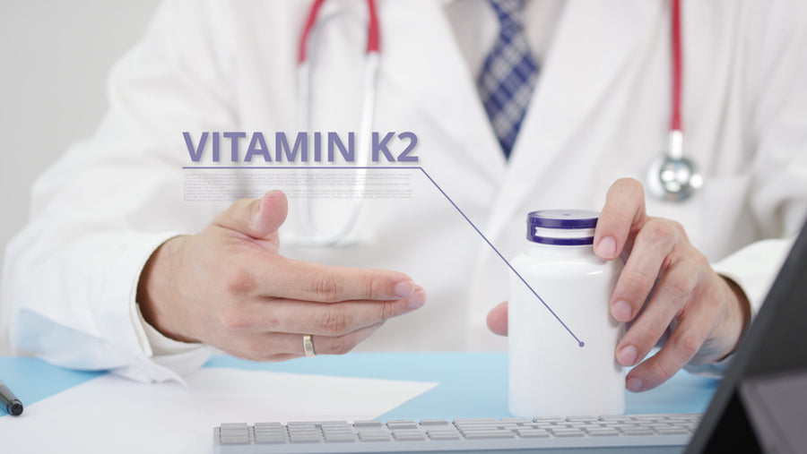 A doctor in a white coat gestures while holding a bottle labeled "Vitamin K2" on a desk with a computer, emphasizing the supplement's importance for health benefits.