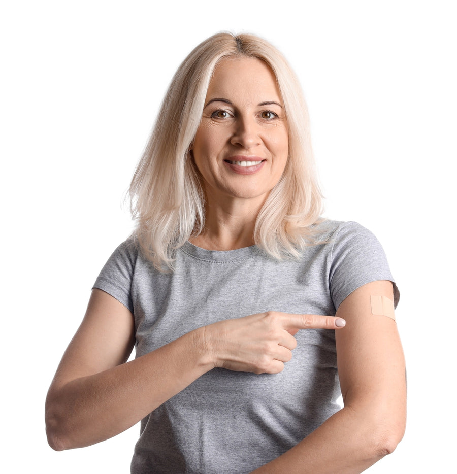 A smiling woman with long blonde hair points to a vitamin patch on her arm while wearing a gray t-shirt, highlighting the benefits of vitamin patches.