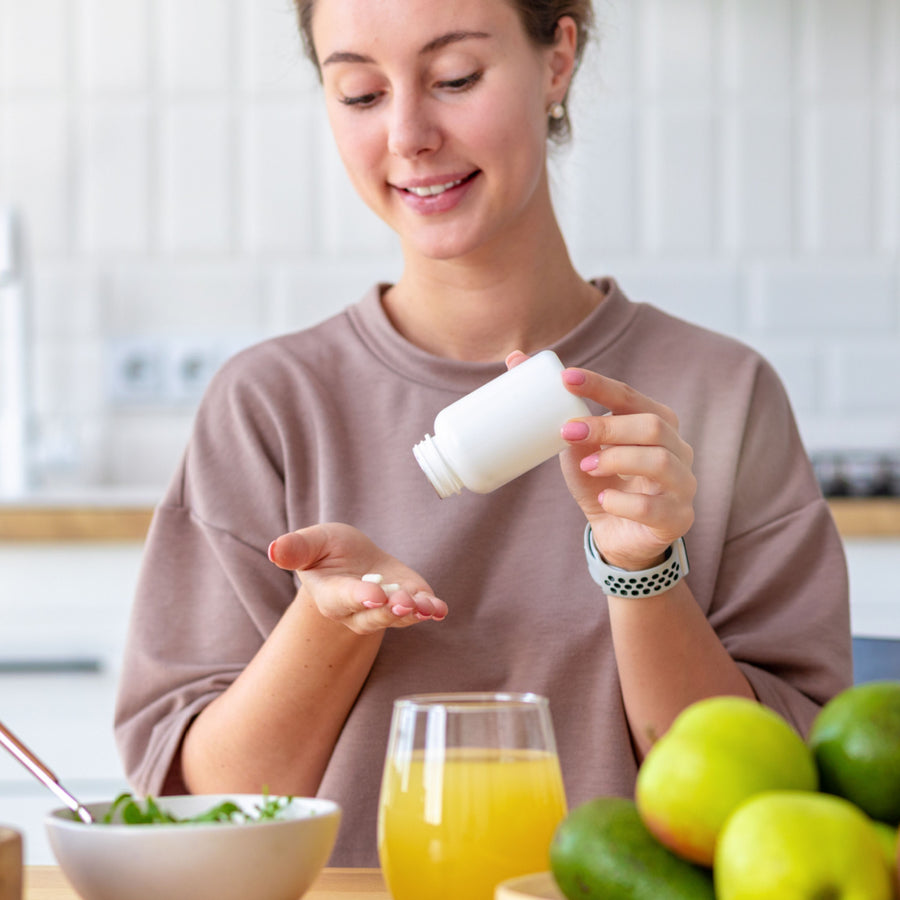 A woman in a cozy kitchen smiles as she pours vitamins from a white bottle into her hand, with a bowl of salad, a glass of orange juice, and fresh fruits on the table nearby.