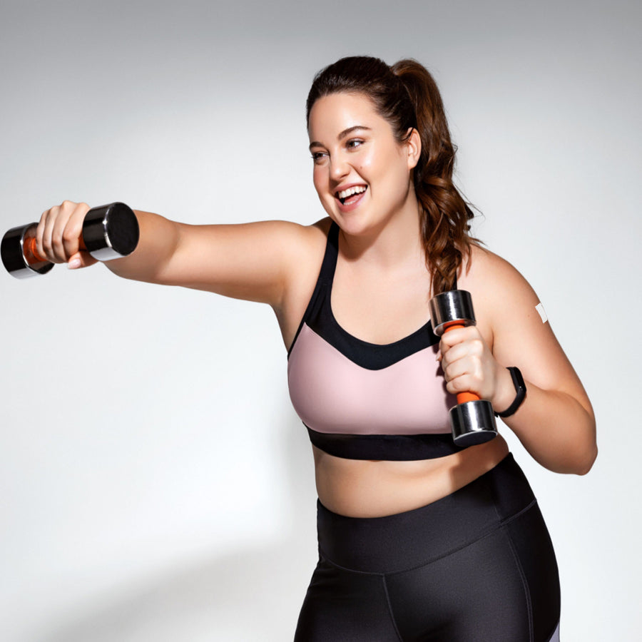A smiling woman in athletic wear holds dumbbells, engaging in a workout against a simple gray background, suggesting an active lifestyle related to weight loss and fitness.