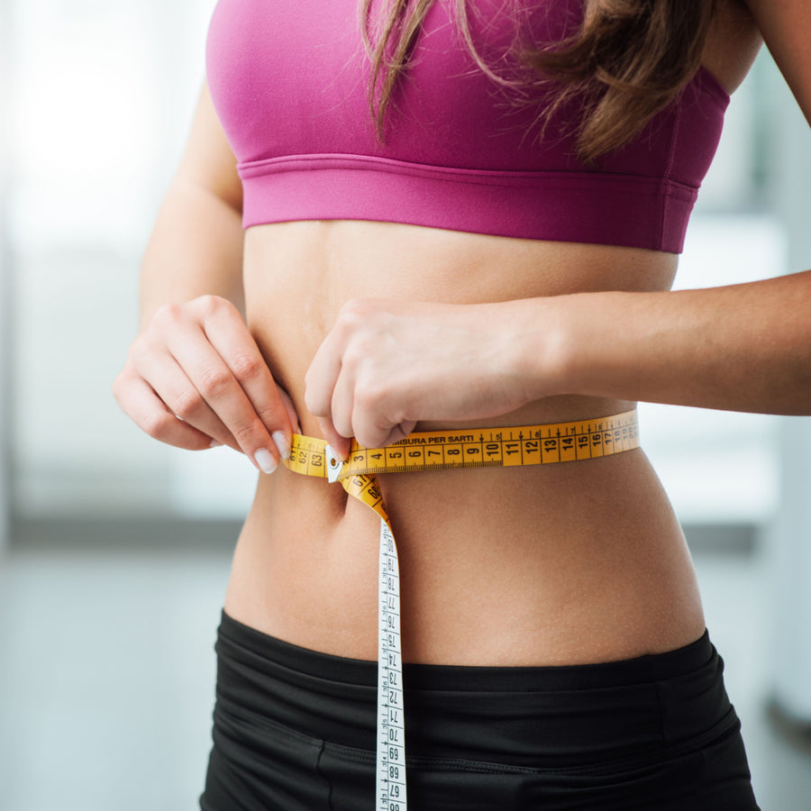 A woman in a pink sports bra measures her waist with a yellow tape measure, focusing intently in a bright, modern indoor setting, emphasizing her commitment to healthy long-term weight loss.