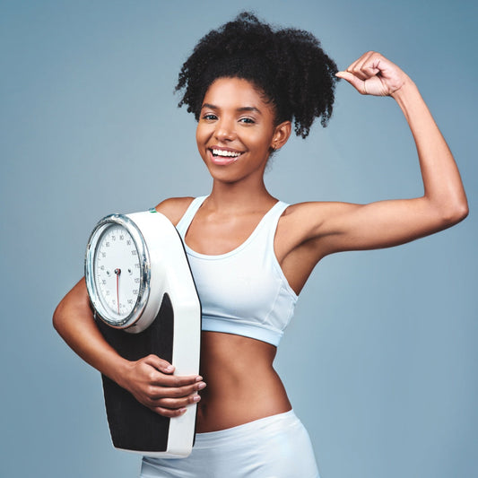 A smiling young woman in a light blue athletic outfit flexes her bicep while holding a weight scale, set against a soft blue background, symbolizing confidence in achieving healthy weight loss.
