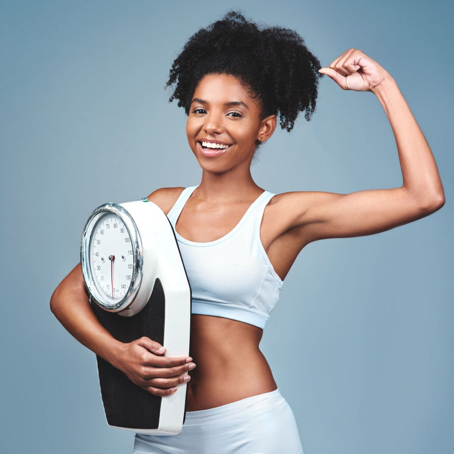A smiling young woman in a light blue athletic outfit flexes her bicep while holding a weight scale, set against a soft blue background, symbolizing confidence in achieving healthy weight loss.